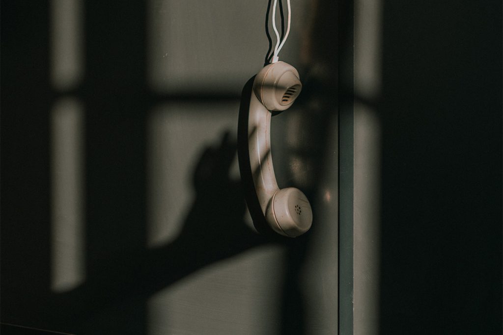corded telephone handset hanging in front of a grey wall, with shadows overlaid of window panes and a hand reaching for the phone.