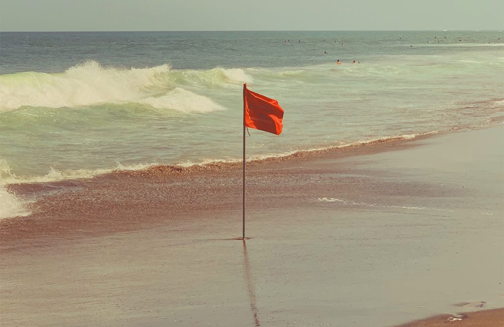 photograph of a red flag planted on the beach with the waves in the background